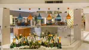 Hall d'hotel moderna con ringhiera in vetro, composizione floreale al centro e lampade pendenti colorate.