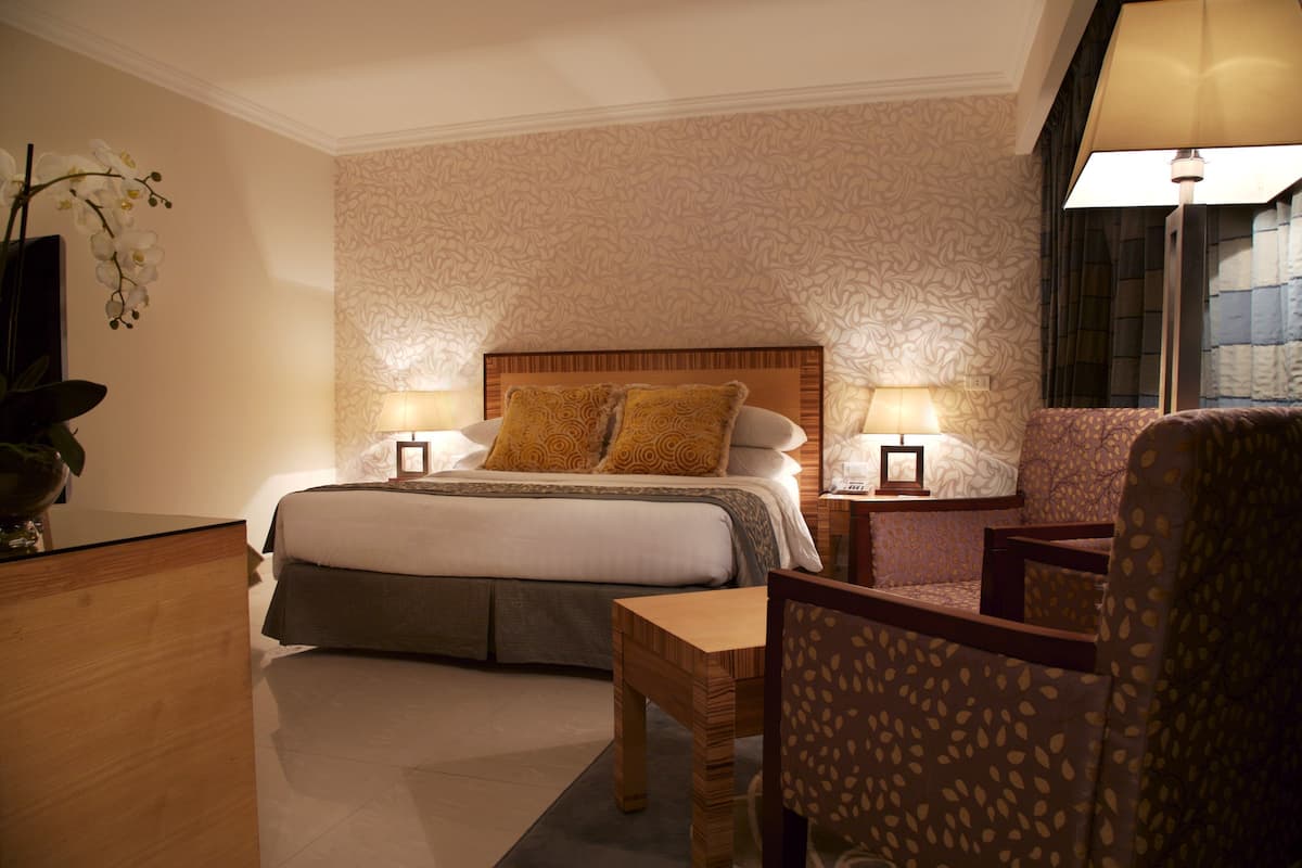 Een hotelkamer met een groot bed, twee nachtkastjes met lampen, een houten bijzettafeltje, een stoel en een decoratieve plant.