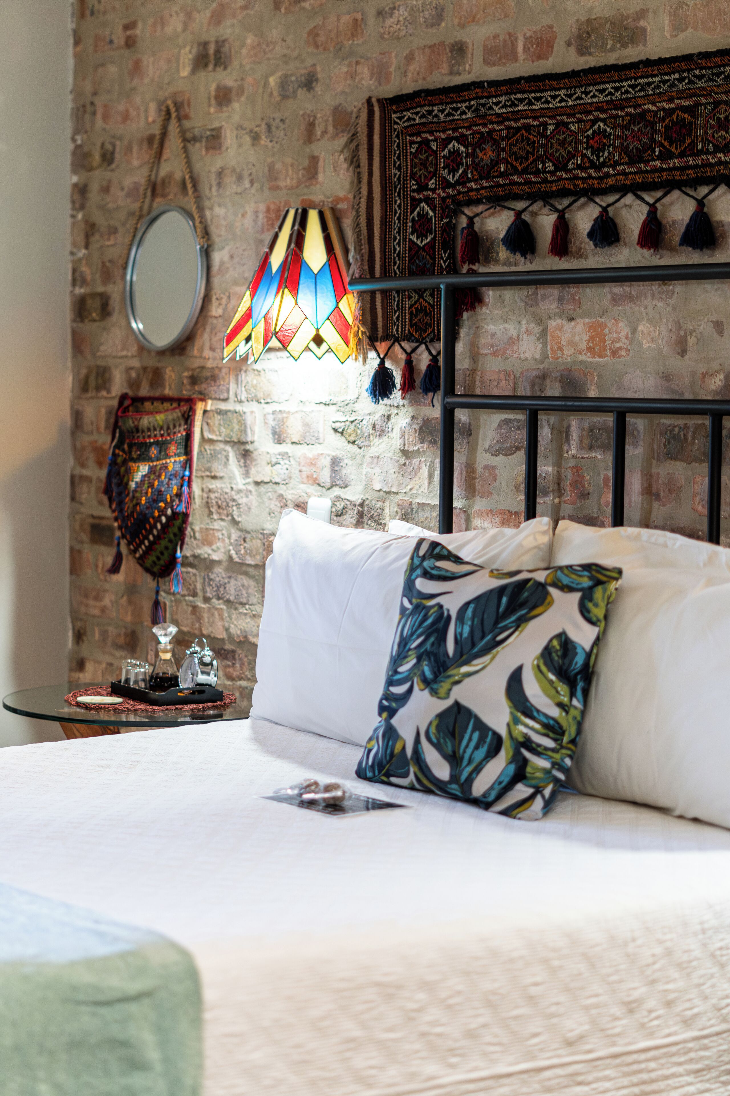 Un dormitorio con pared de ladrillo, una cama con ropa blanca, una lámpara colorida y un tapiz colgado.