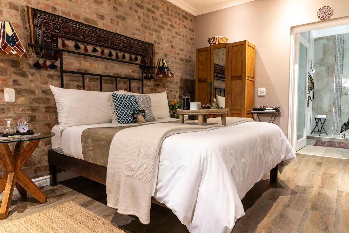A bedroom with a brick wall, a bed with white bedding, a wooden headboard, and a small table with a lamp.