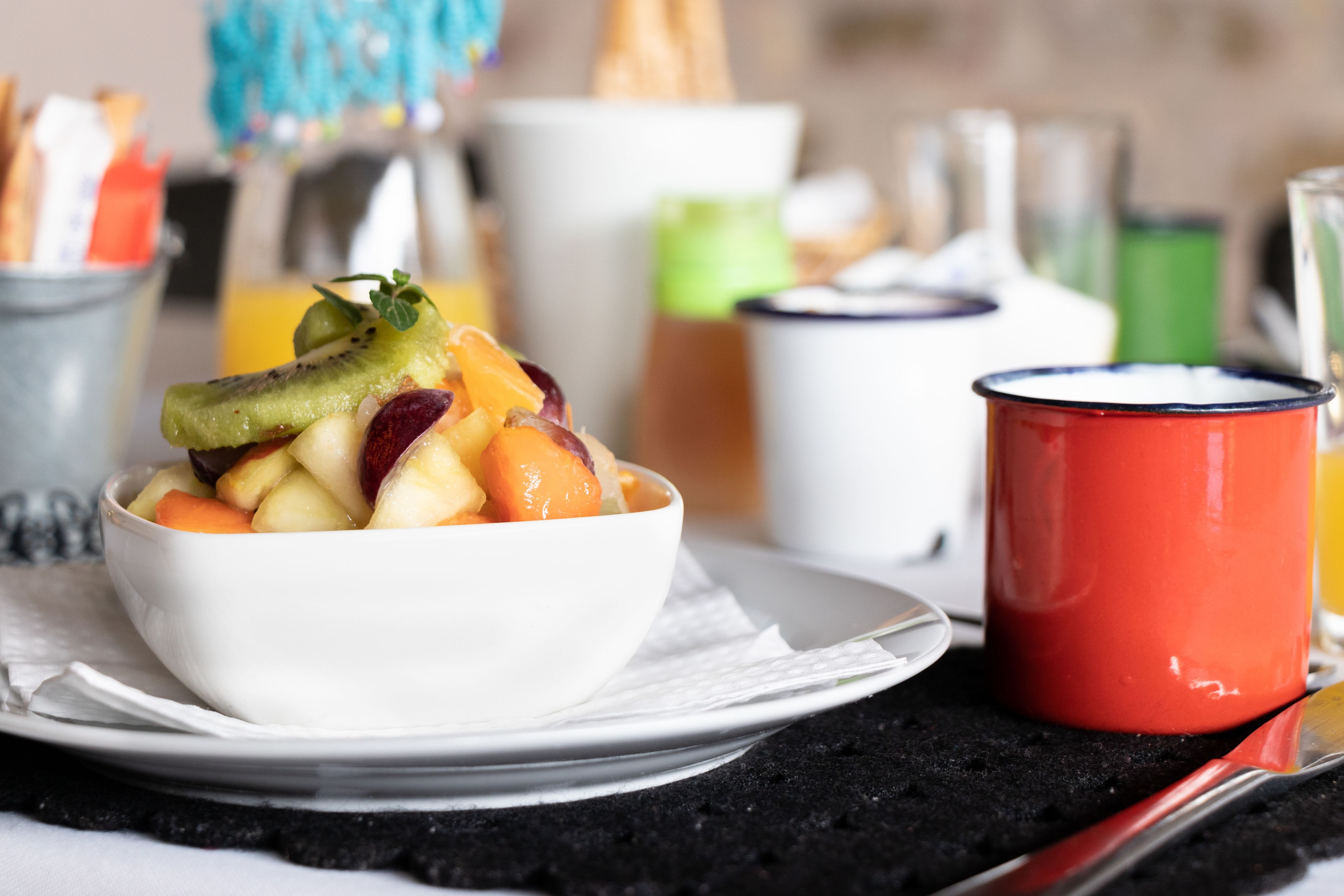 A white bowl of fruit on a plate with a red mug beside it.