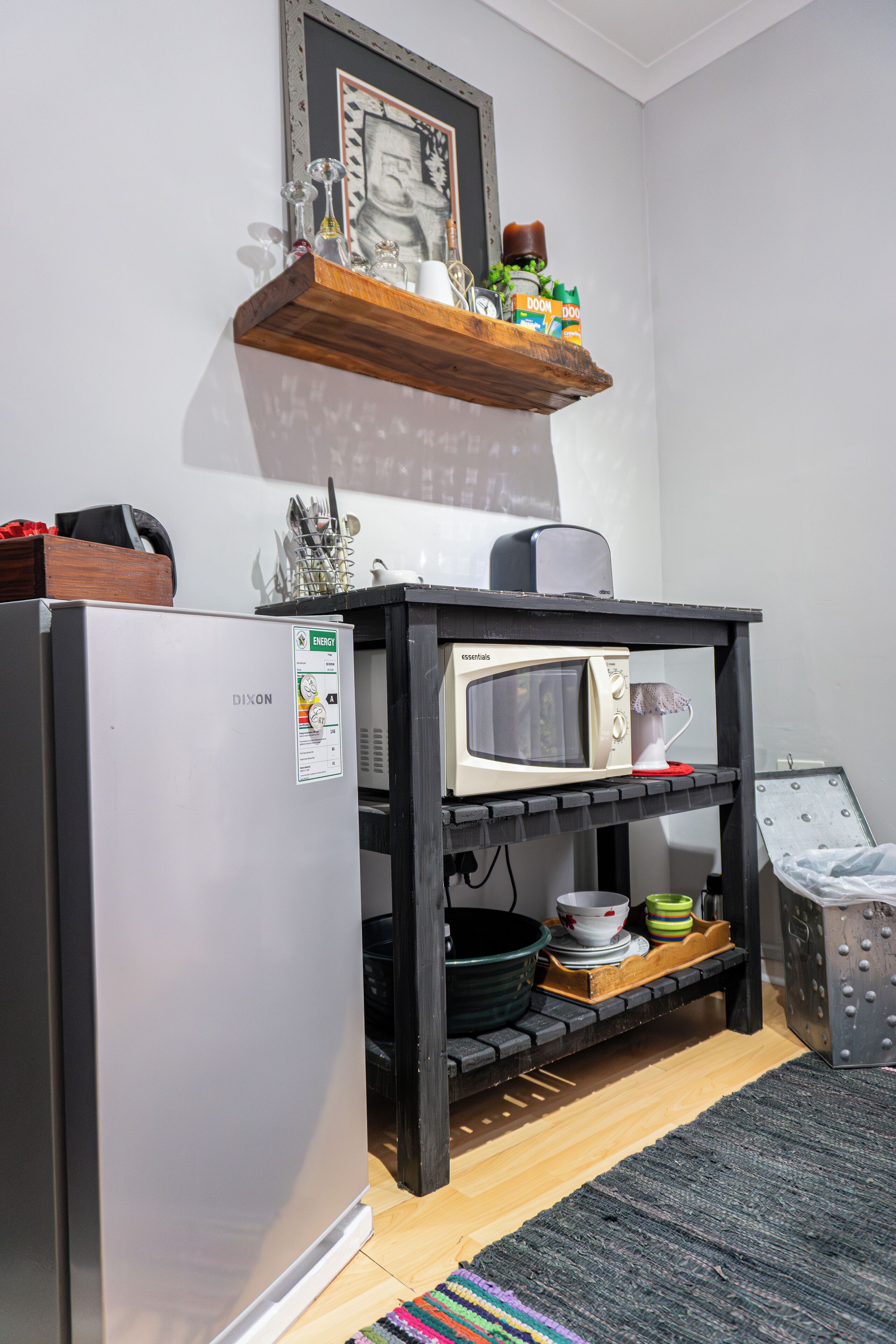 A compact kitchen with a refrigerator, microwave, and a wooden shelf above holding various kitchen items.