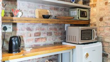 Une cuisine compacte avec un mur en briques, des étagères en bois, un four à micro-ondes, un petit réfrigérateur et un panier en osier.