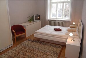 Ein Schlafzimmer mit einem Bett, einem roten Sessel, einem kleinen Fernseher und einem Fenster mit Blick auf Bäume.