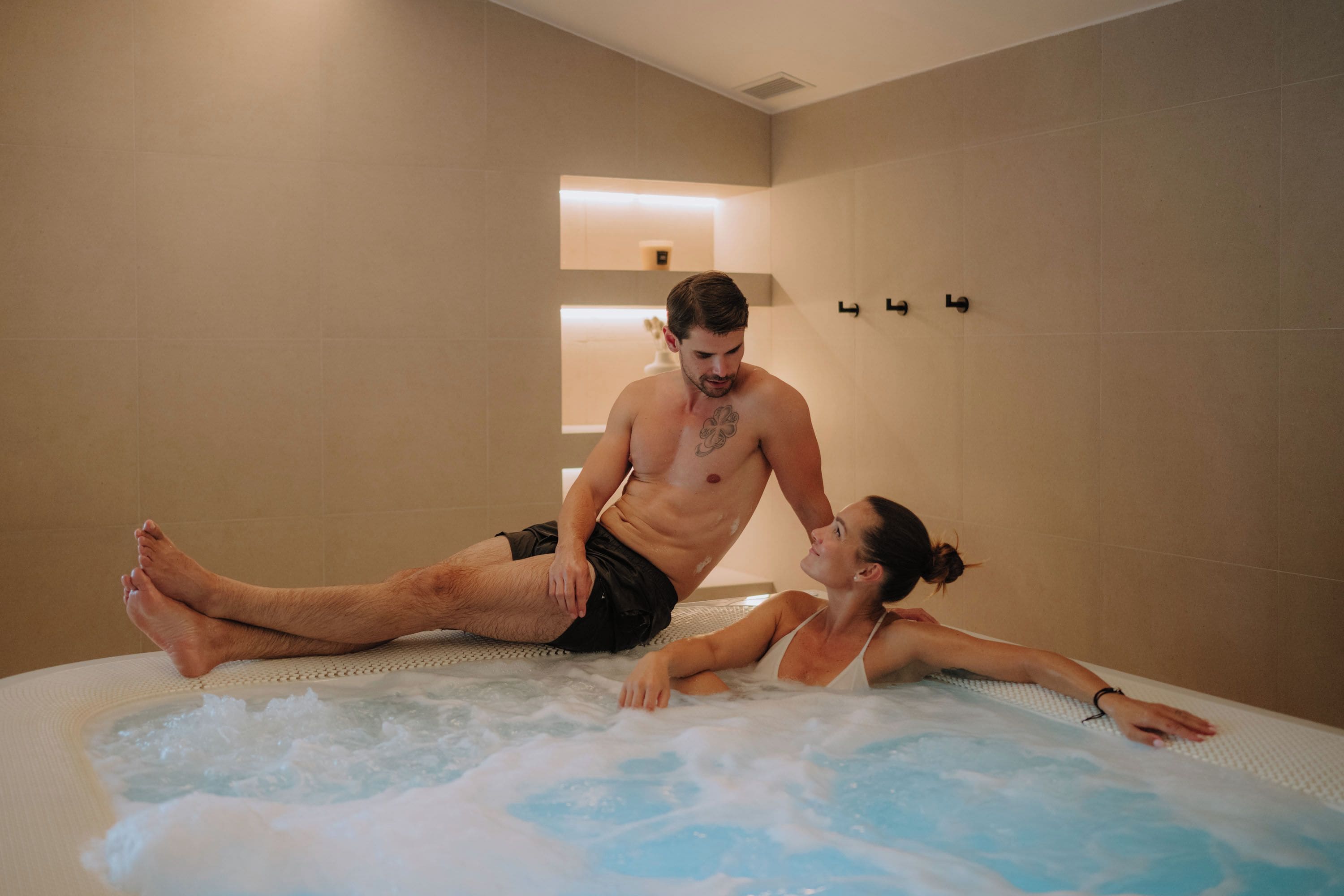 A couple in a hot tub with a tiled wall and a small shelf in the background.