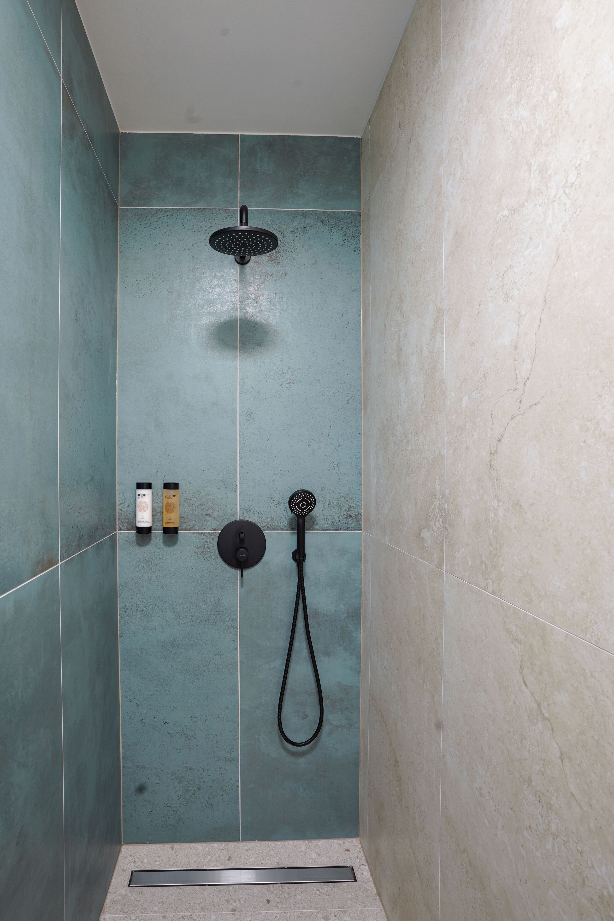A shower area with a handheld showerhead and a wall-mounted showerhead. There are two bottles of shampoo and conditioner on the wall.