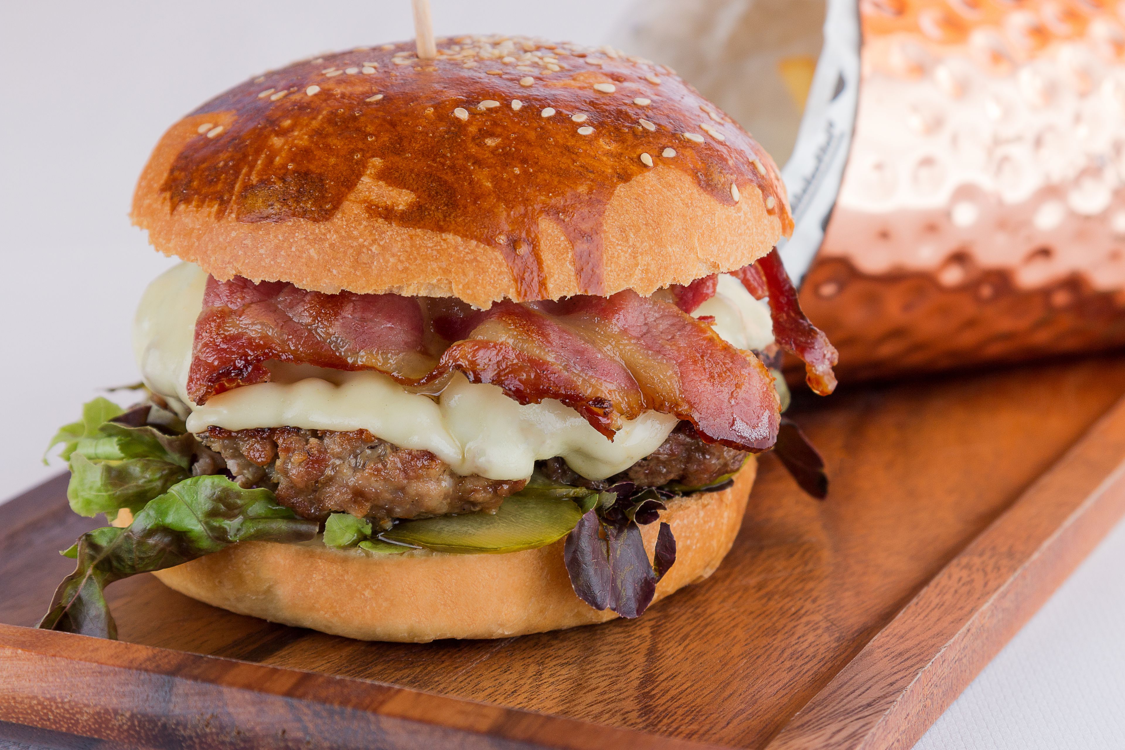 A burger with lettuce, pickles, bacon, and cheese on a sesame seed bun.