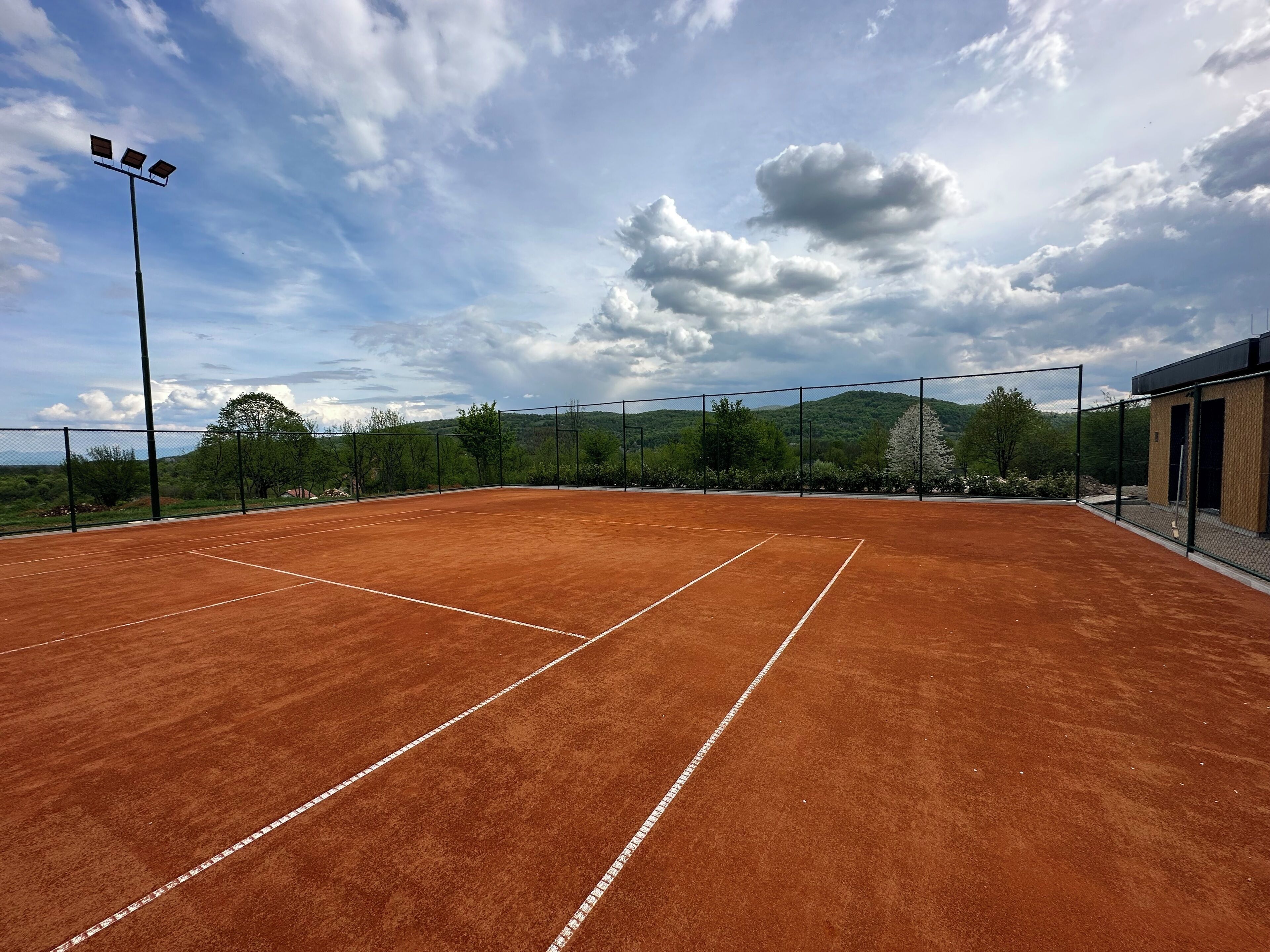 An outdoor tennis court with a net, surrounded by a fence, and a building with a wooden exterior.