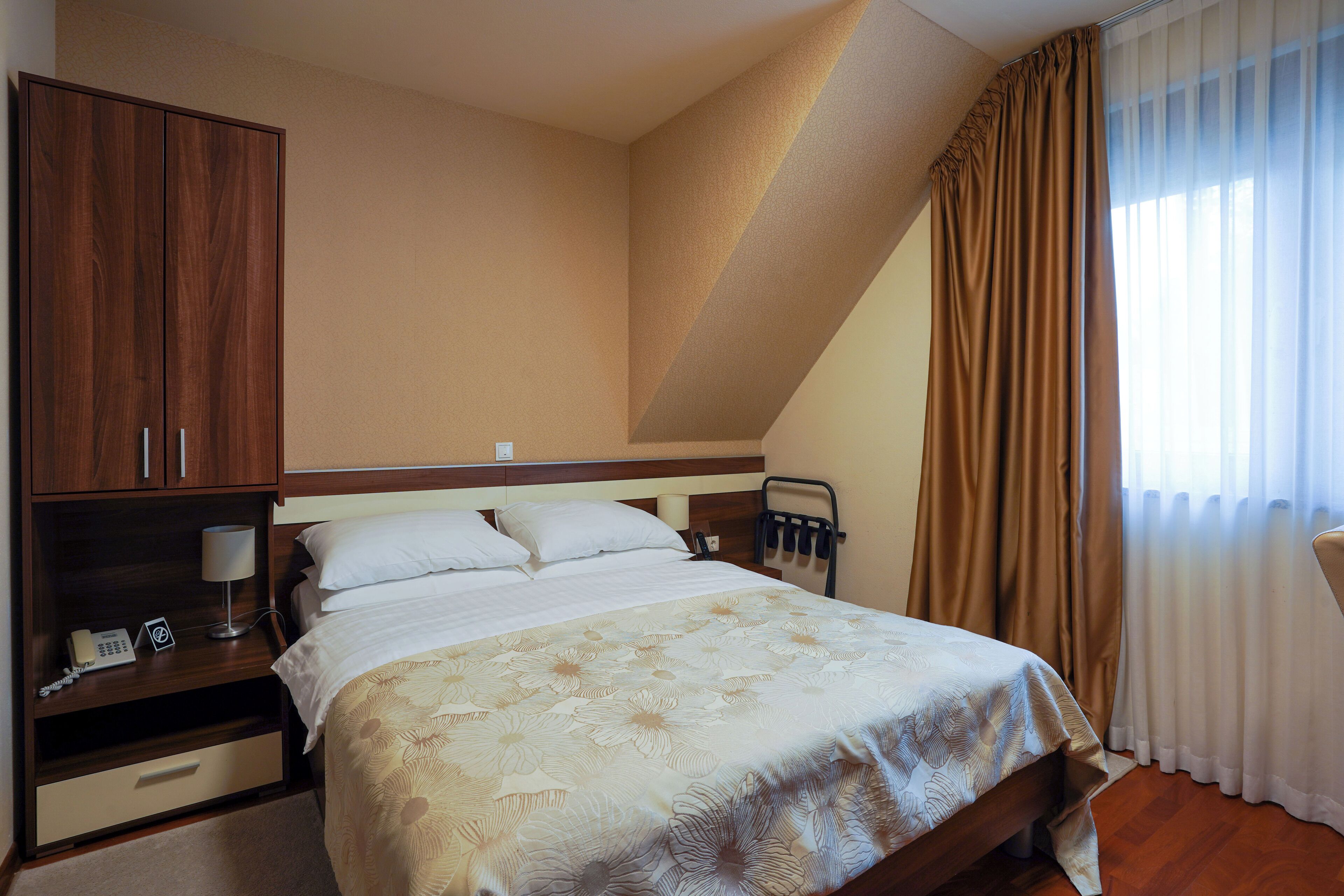 A hotel room with a bed, a nightstand with a lamp, a wooden wardrobe, and a window with curtains.