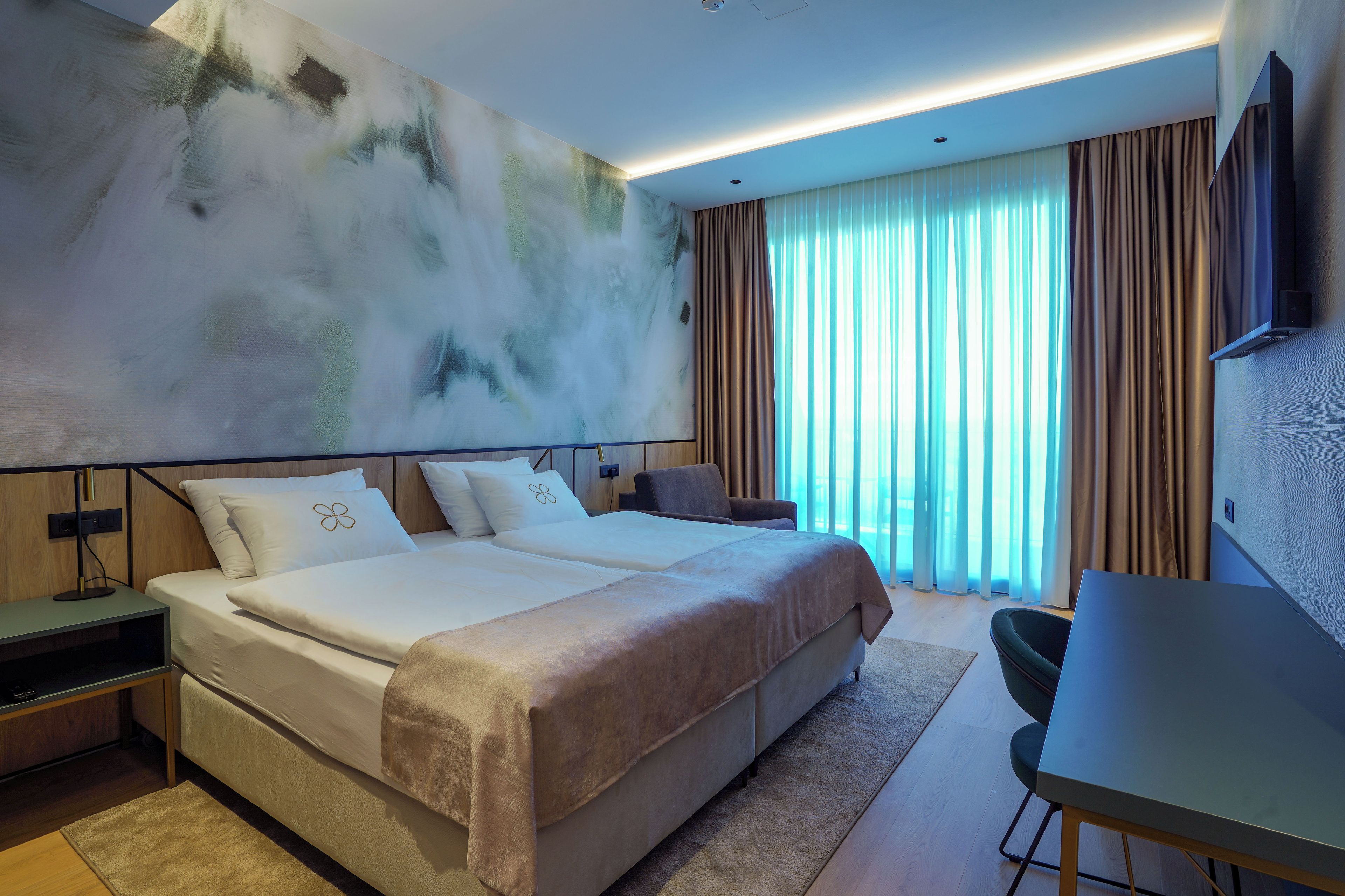 A modern hotel room with a large bed, a desk, and a view of the outdoors through sheer curtains.
