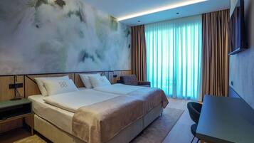 A modern hotel room with a large bed, a desk, and a view of the outdoors through sheer curtains.