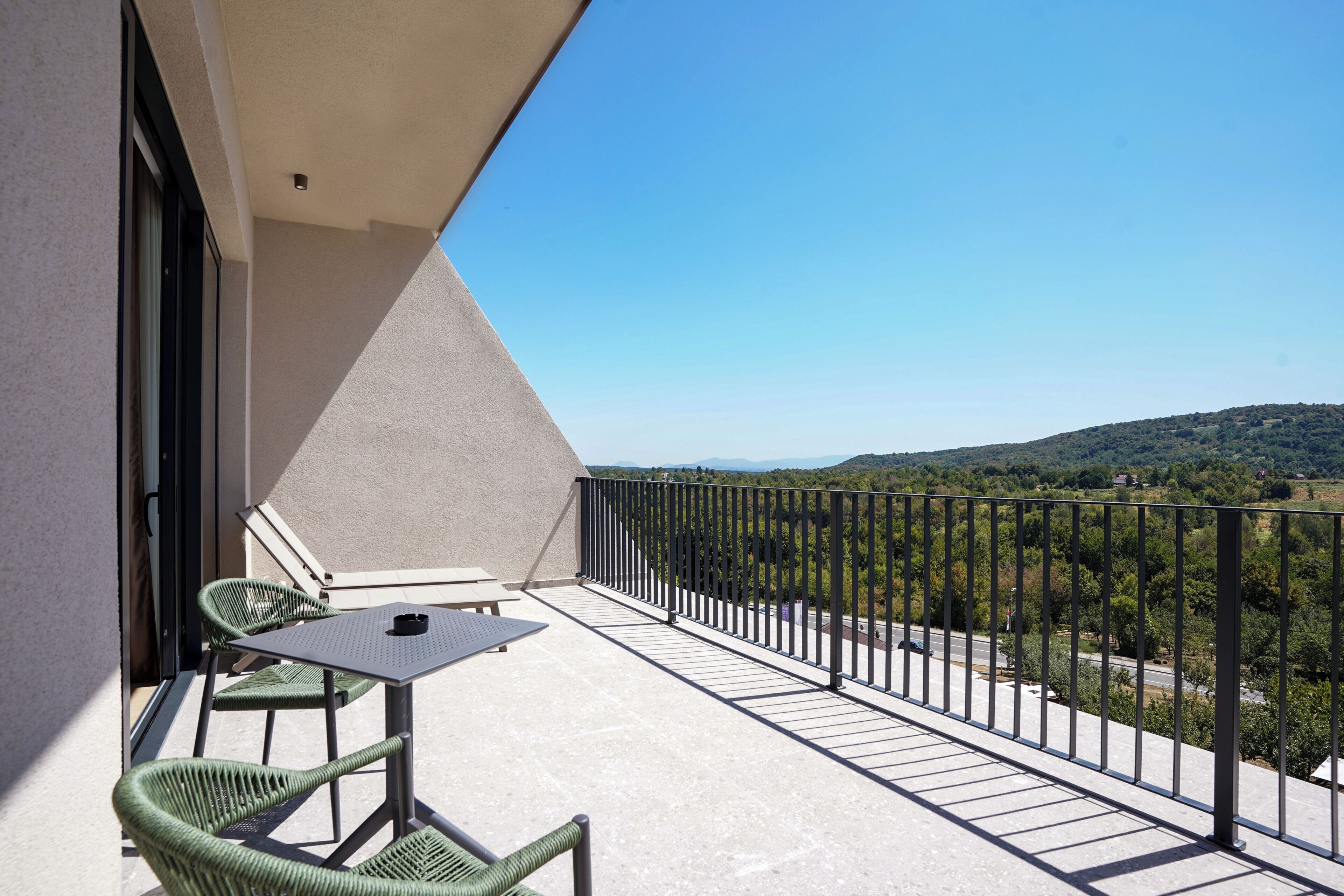 A balcony with a table and chairs, a view of a road and greenery, and a clear sky.