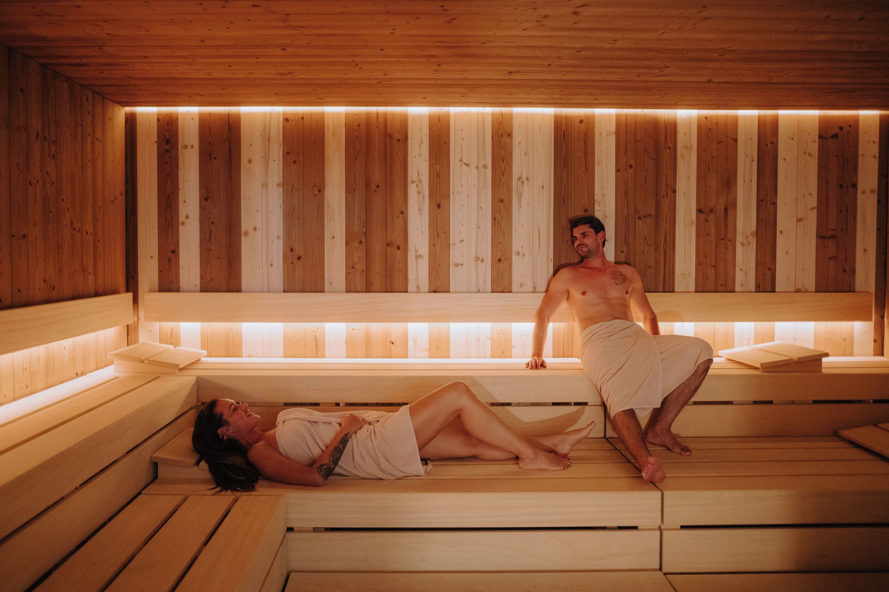 A sauna with wooden benches and benches, a person sitting on a bench, and another person lying on a bench.