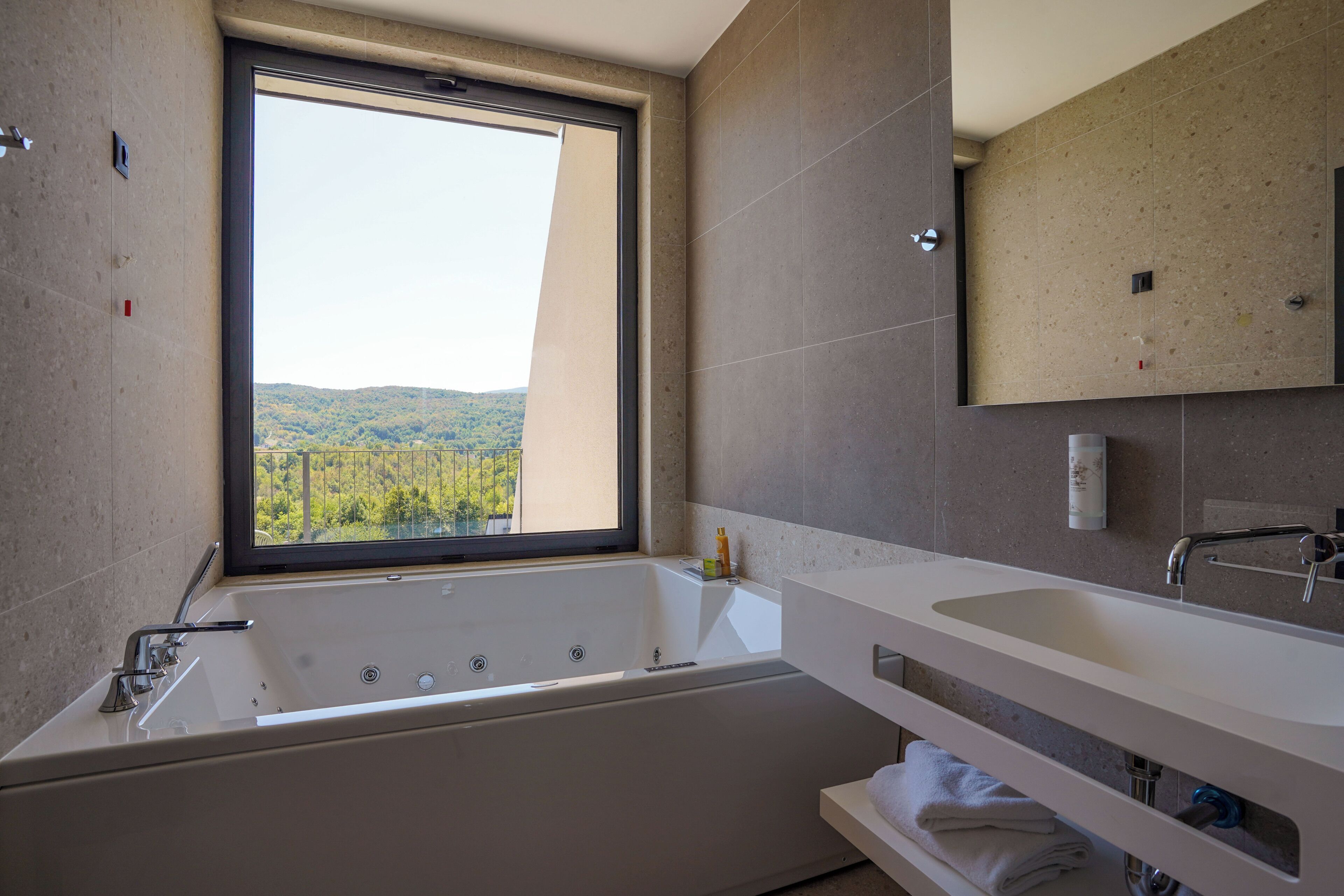 Ein modernes Badezimmer mit einer großzügigen Badewanne, einem Fenster mit Blick auf Grünflächen und einem Spiegel über dem Waschbecken.
