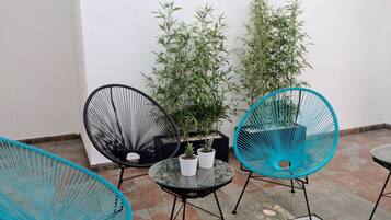 En liten sittplats utomhus med två turkosa stolar och ett bord, omgiven av planterade växter och en vit vägg.
