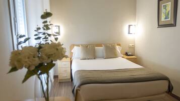 Una habitación de hotel con cama, mesitas de noche, un jarrón con flores y un teléfono en la pared.
