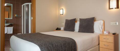 Quarto de hotel com uma cama grande, mesa de cabeceira de madeira com um telefone e um aviso, luz acoplada à parede e banheiro visível por uma porta aberta.