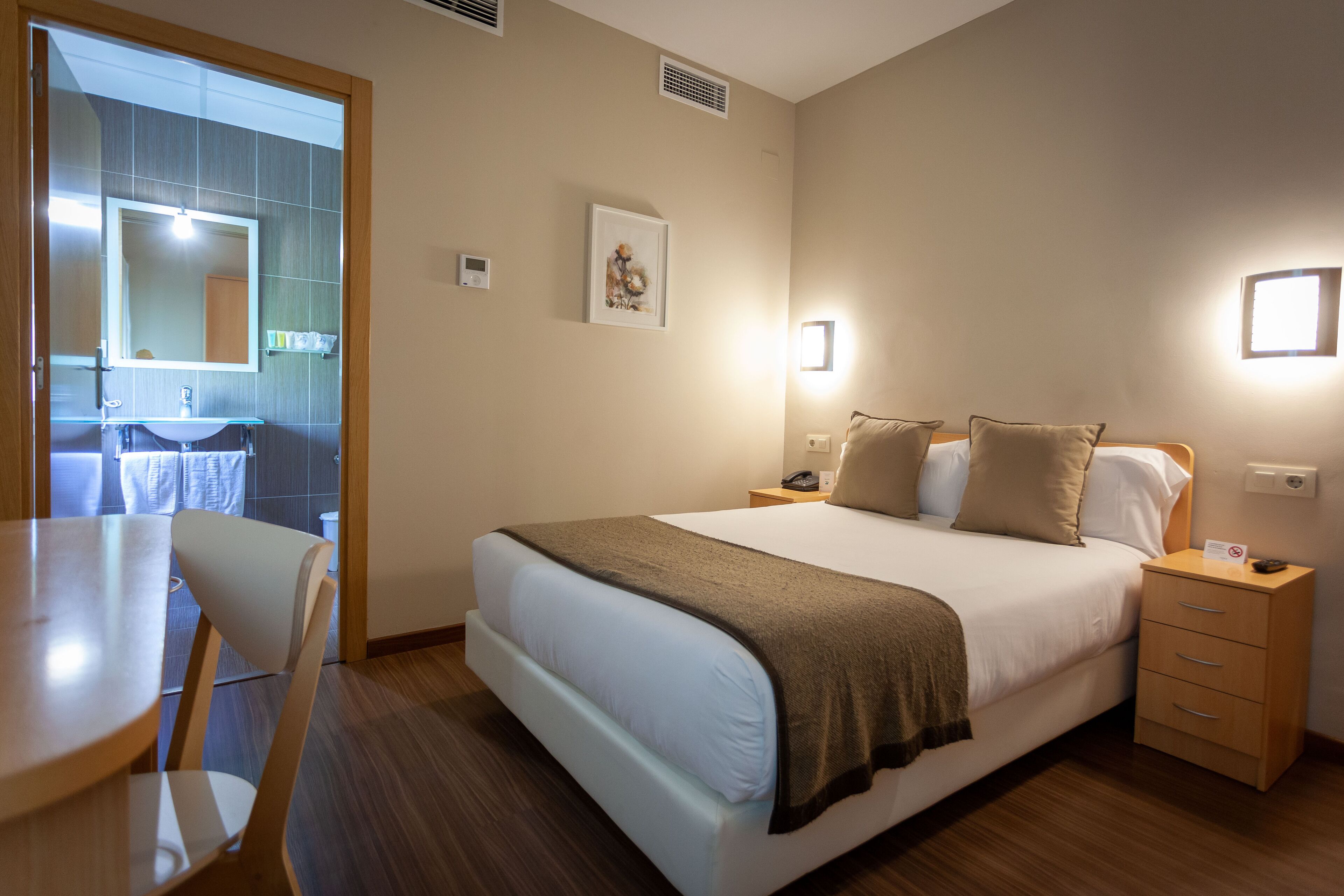 Habitación de hotel con cama, mesita de noche y una silla. Se ve un baño por una puerta abierta.