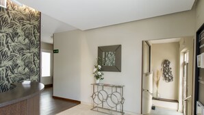 A modern reception area with a wooden reception desk, a wall-mounted mirror, and a minimalist console table with a vase of flowers. - Hotel Vettonia (Merida)