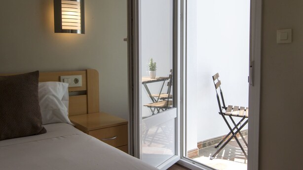 A bedroom with a bed, a bedside table, a wall-mounted light, and a sliding glass door leading to a balcony with a chair and a small table. - Hotel Vettonia (Merida)