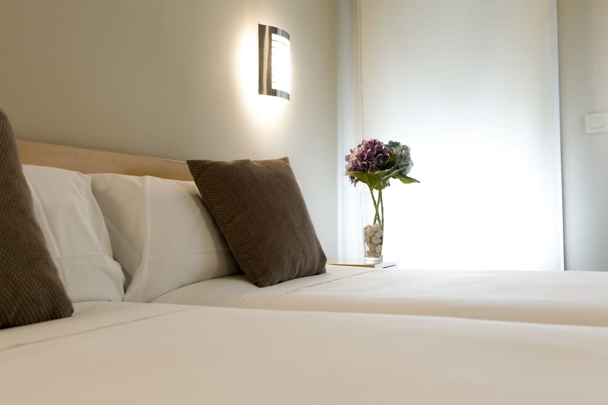 A neatly made bed with white and brown pillows, a bedside table with a vase of flowers, and a wall-mounted light fixture.