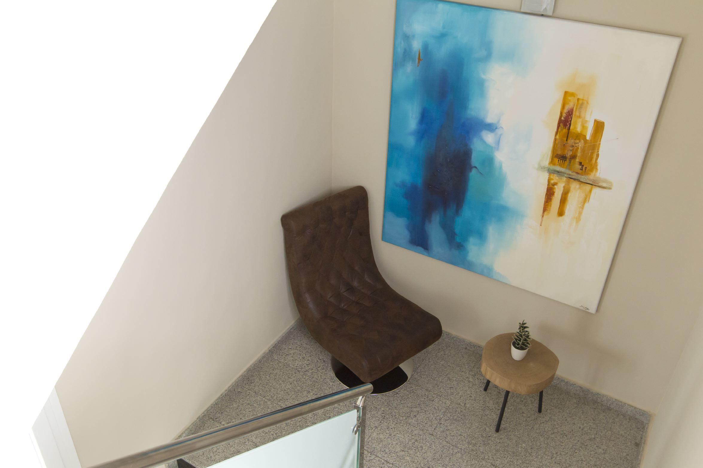 Un interior moderno con una butaca marrón curvada, una pequeña mesa redonda con una planta en maceta y un cuadro abstracto grande en la pared.