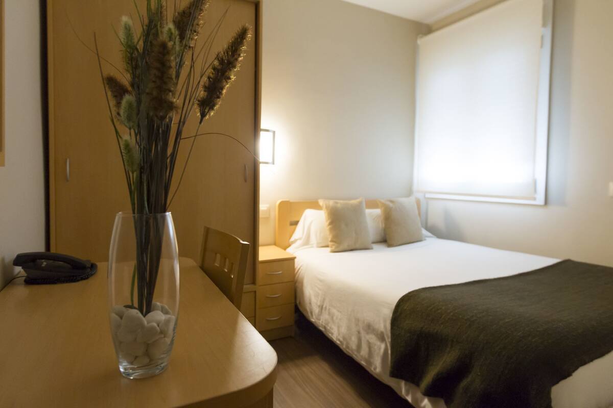 A hotel room with a bed, bedside table, and a vase with dried plants.