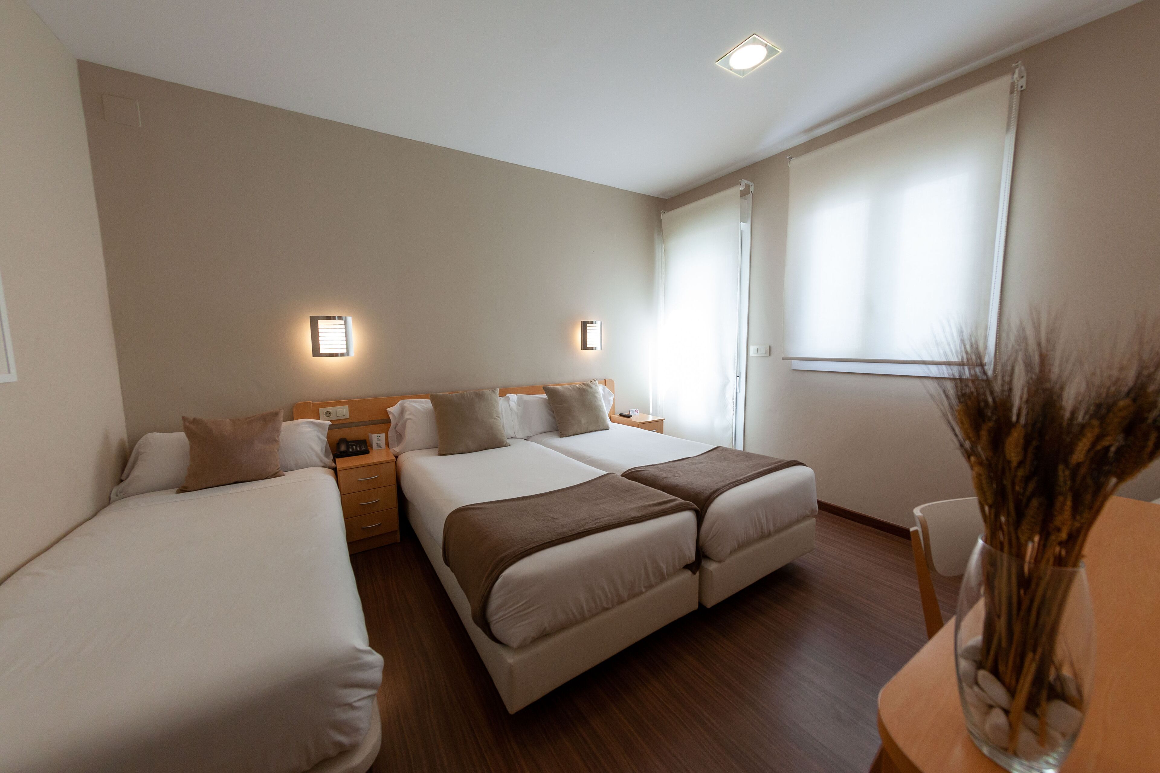 Une chambre d’hôtel avec deux lits simples, un lit double, une table de chevet en bois, une petite table avec un vase et une fenêtre avec des stores.