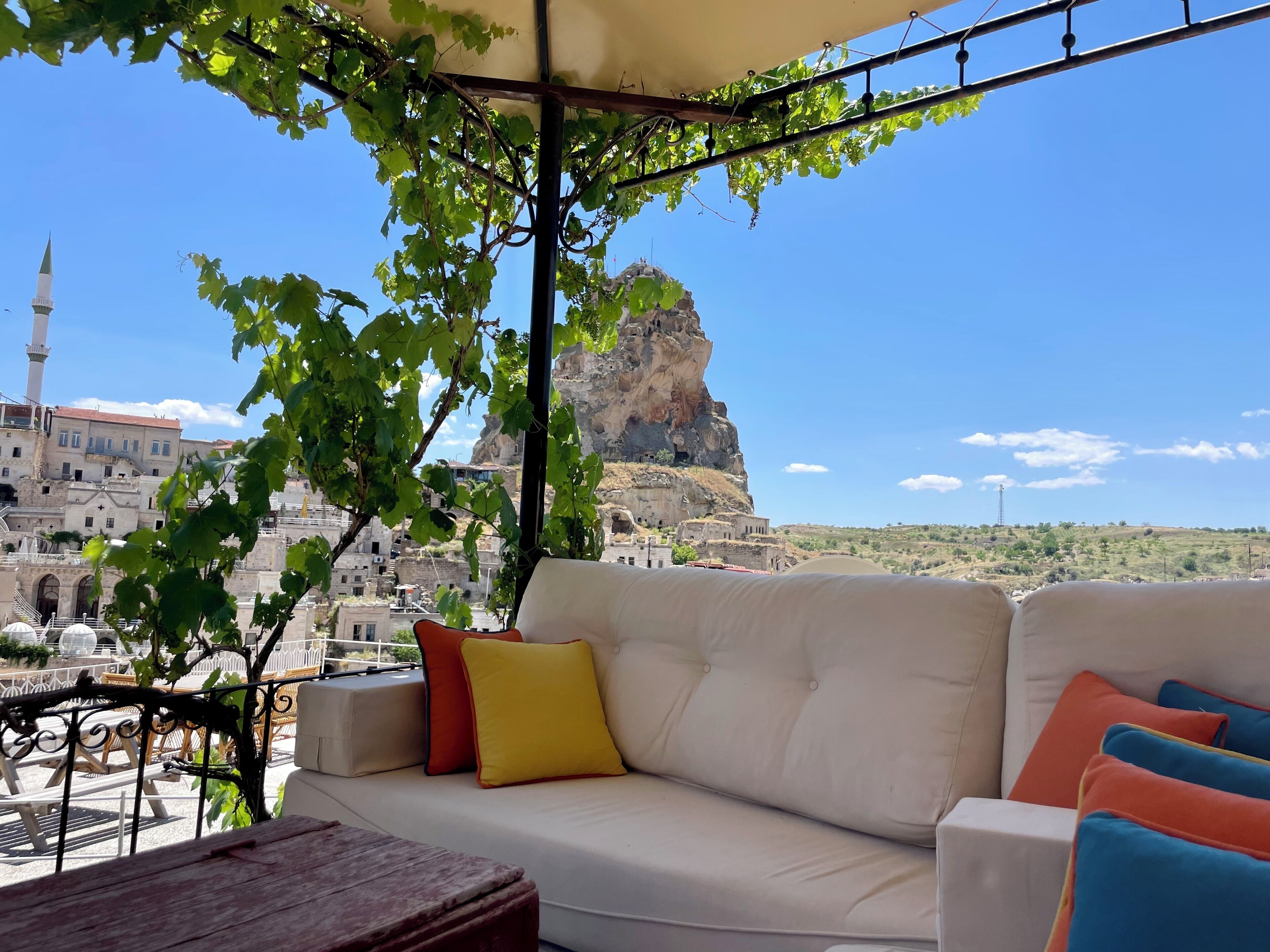 A balcony with a white sofa, colorful cushions, and a view of a historic building and a rocky cliff.