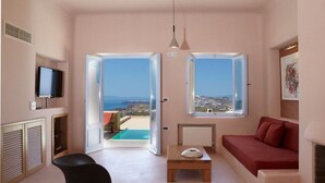 Una sala moderna con un sofá rojo, un reposapiés negro y una mesa de centro de madera. Hay dos ventanas grandes que ofrecen vistas al mar y un balcón.