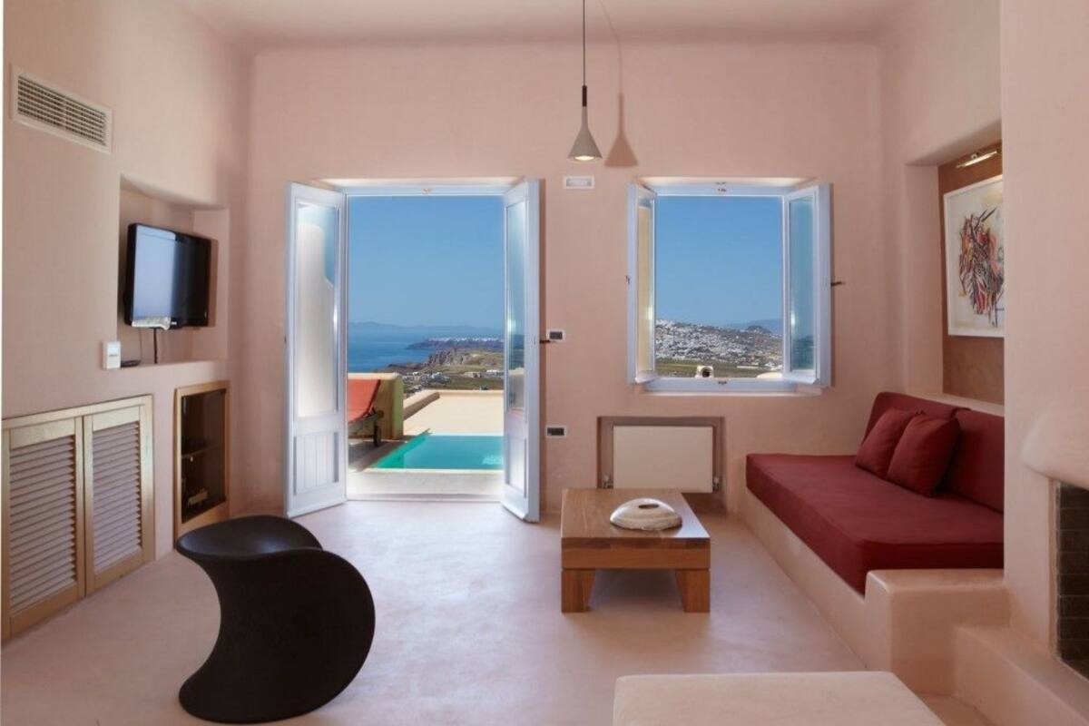 Ein modernes Wohnzimmer mit einem roten Sofa, einem schwarzen Ottomanen und einem Holztisch. Zwei große Fenster bieten Blick auf das Meer und es gibt einen Balkon.