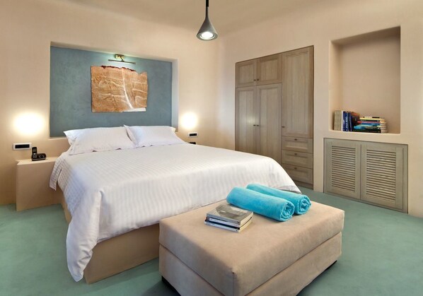 A bedroom with a bed, bedside tables, a nightstand, a wall-mounted artwork, a wardrobe, and a bookshelf. - Voreina Gallery Suites (Santorini)
