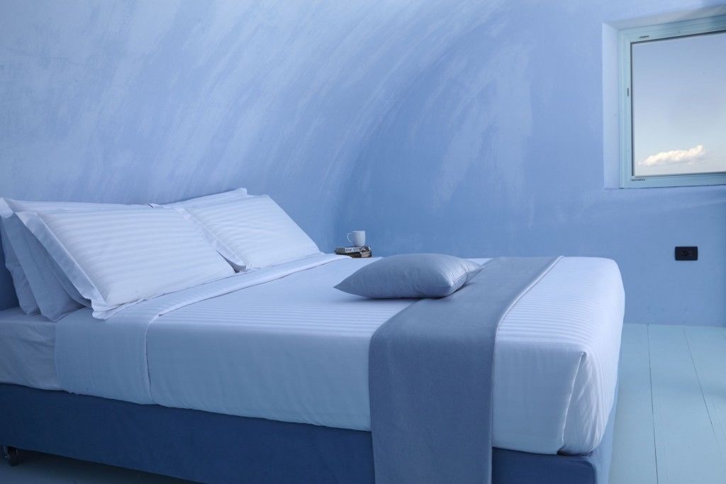 A bed with white linens and a grey pillow in a room with light blue walls and a window.