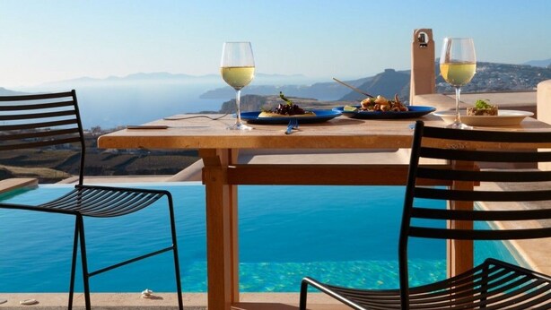Una mesa de comedor con dos platos de comida y dos copas de vino blanco, con vista a una piscina azul cristalina y un paisaje pintoresco de montañas y el mar.