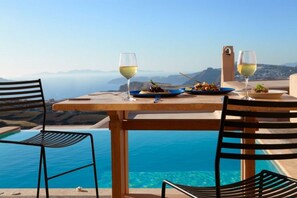 A dining table with two plates of food and two glasses of white wine, overlooking a clear blue pool and a scenic view of mountains and the sea. - Voreina Gallery Suites (Santorini)