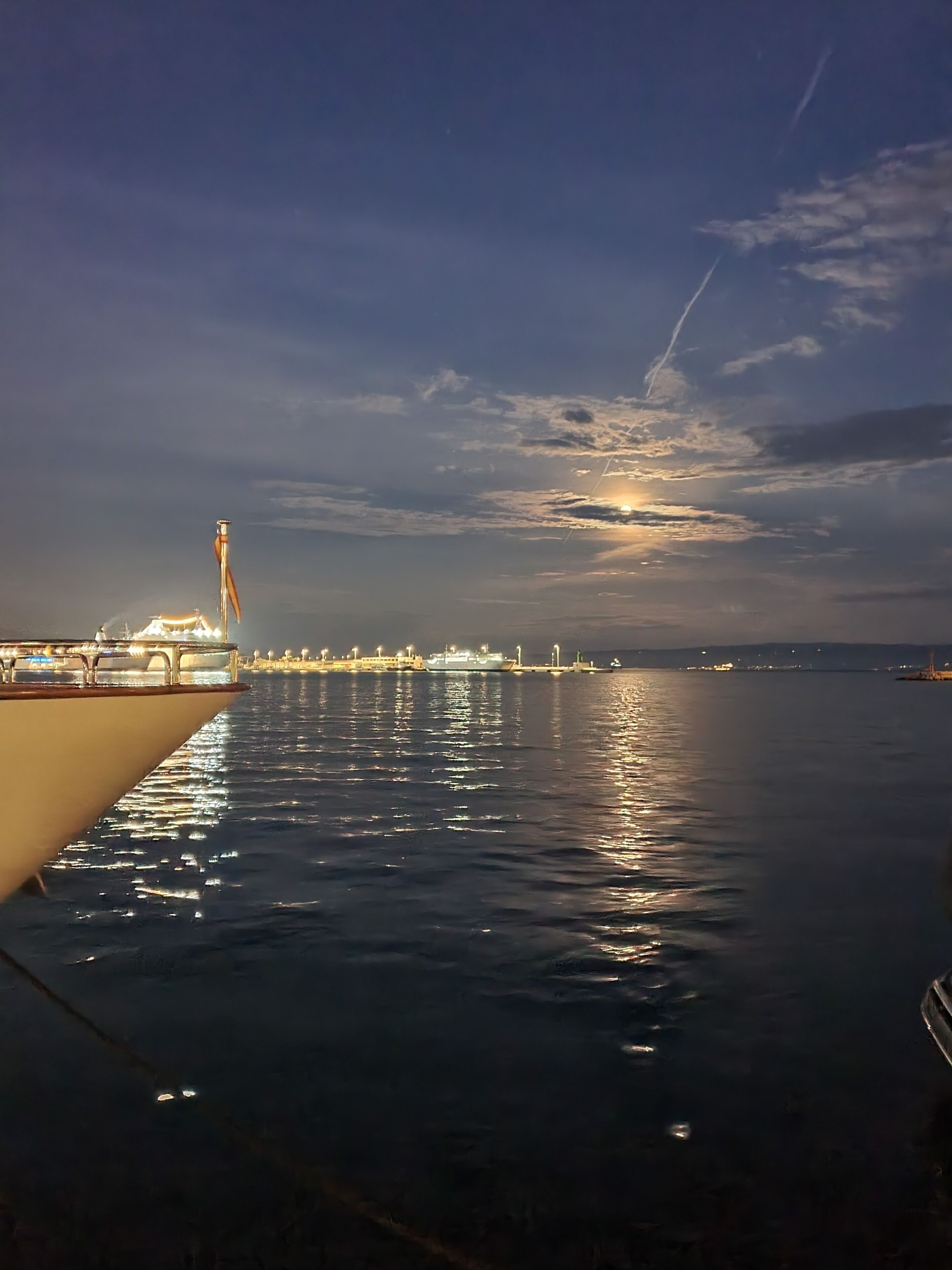 A calm waterfront scene with a large ship docked at a pier, reflecting the sky and lights from the ship and surrounding area.