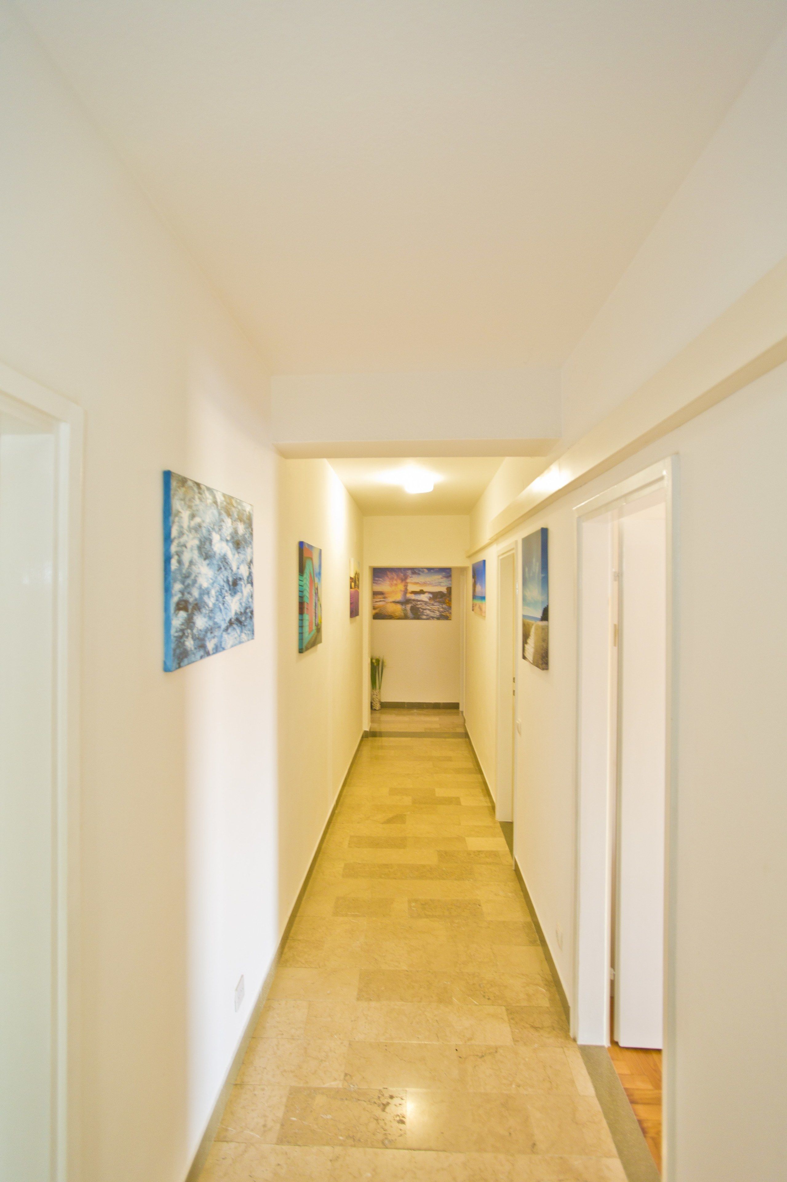A narrow hallway with white walls and a wooden floor. There are several doors on the right side and artwork on the left.