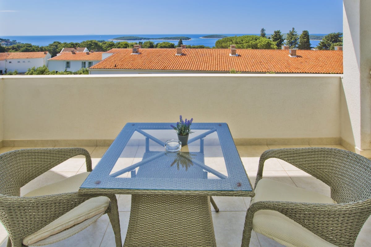 A balcony with a glass table and wicker chairs, overlooking a coastal area with terracotta roofs and greenery.
