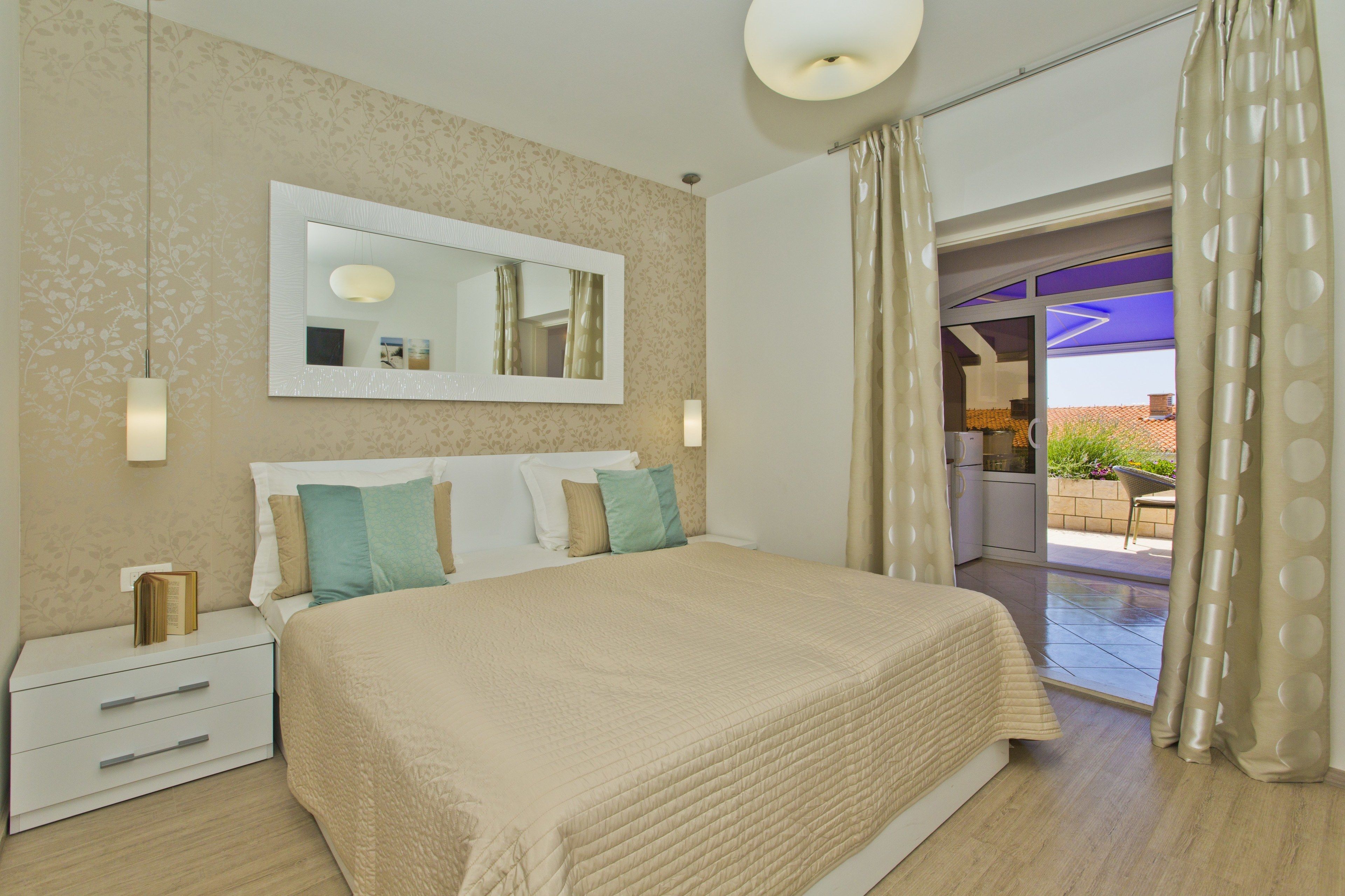 A bedroom with a large bed, a nightstand, a mirror, and a view of the outside.