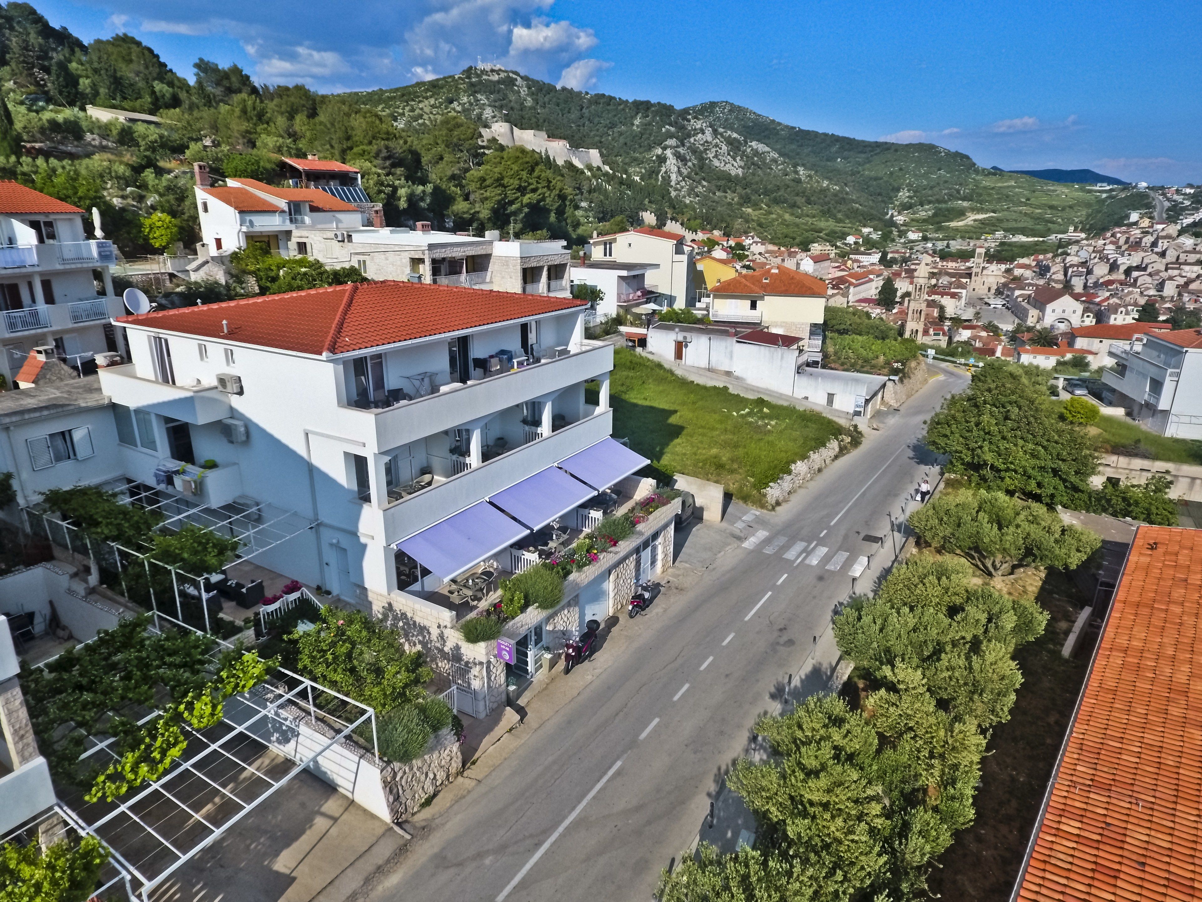 A multi-story building with balconies, a street in the foreground, and a mountainous backdrop.