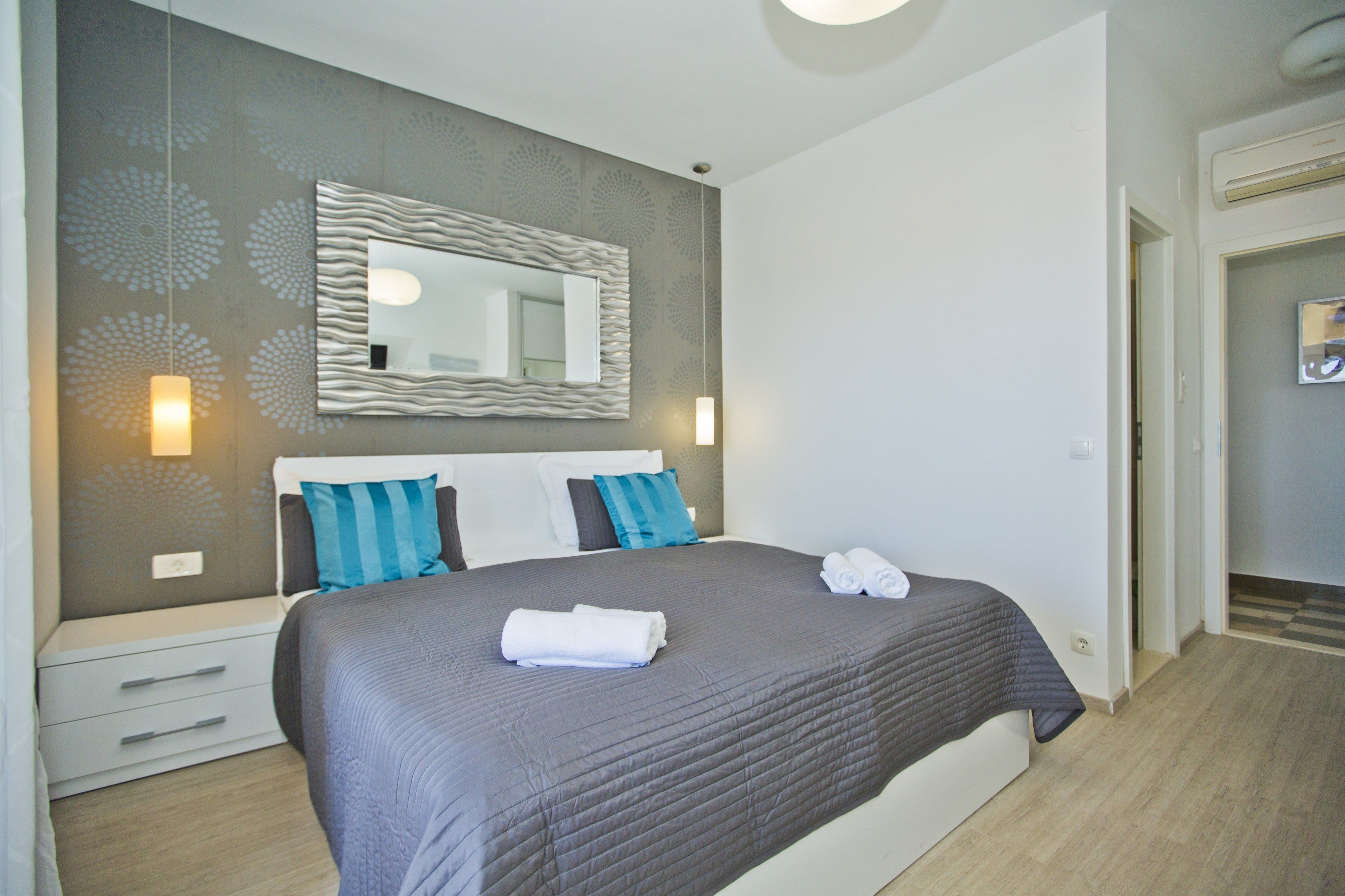A modern bedroom with a large bed, a wall-mounted mirror, bedside lamps, and a neatly made bed with towels.