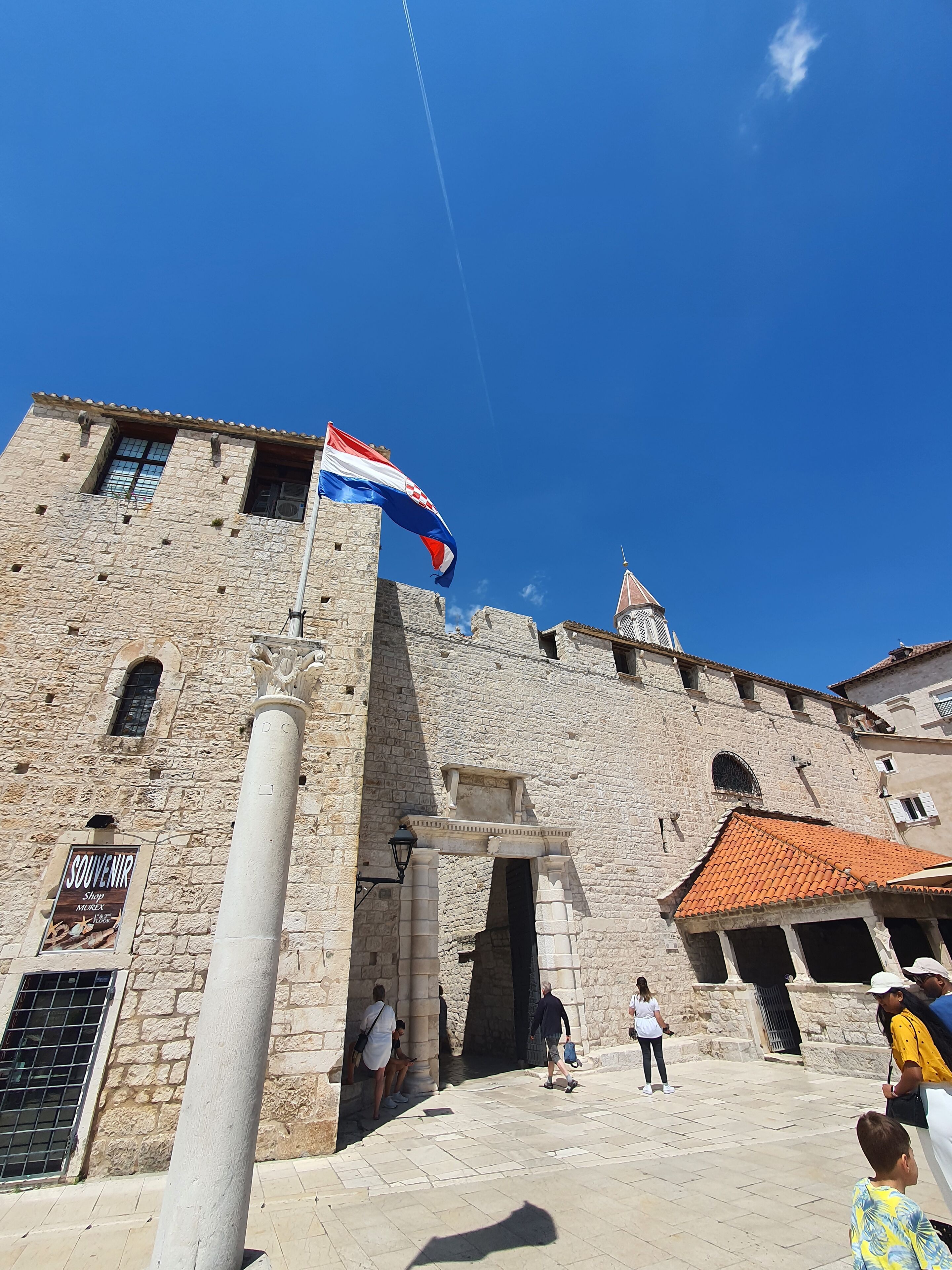 A historic stone building with a flag, a central entrance, and people around.