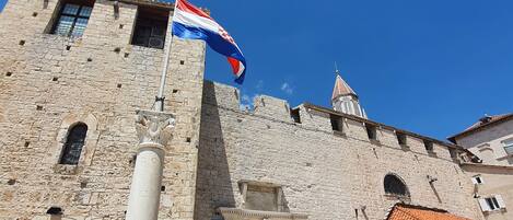 A historic stone building with a flag, a central entrance, and people around.