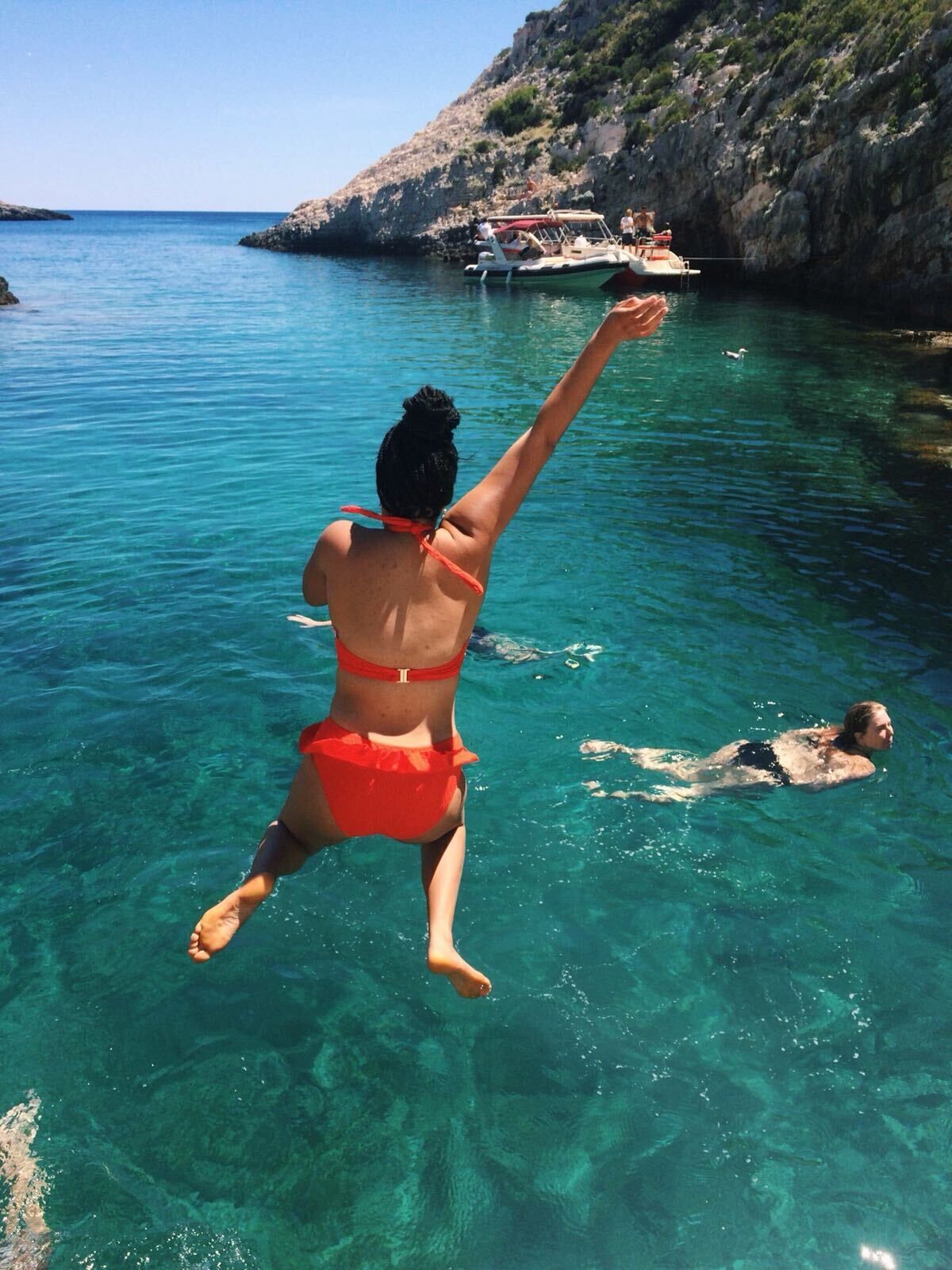 A person in a red bikini diving into clear blue water with a rocky coastline and a boat in the background.