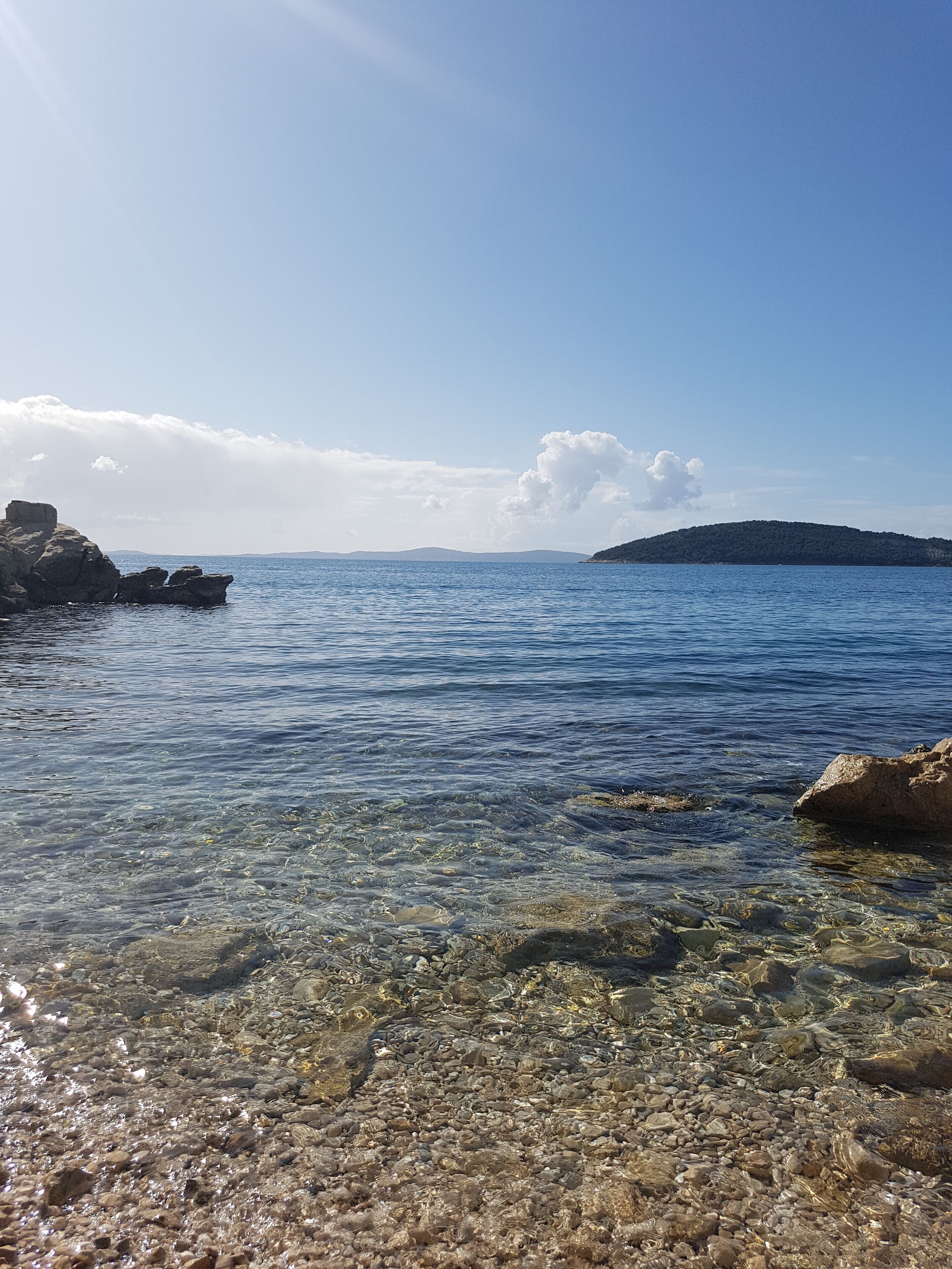 A clear view of a rocky shoreline with calm water, a distant hill, and a partly cloudy sky.