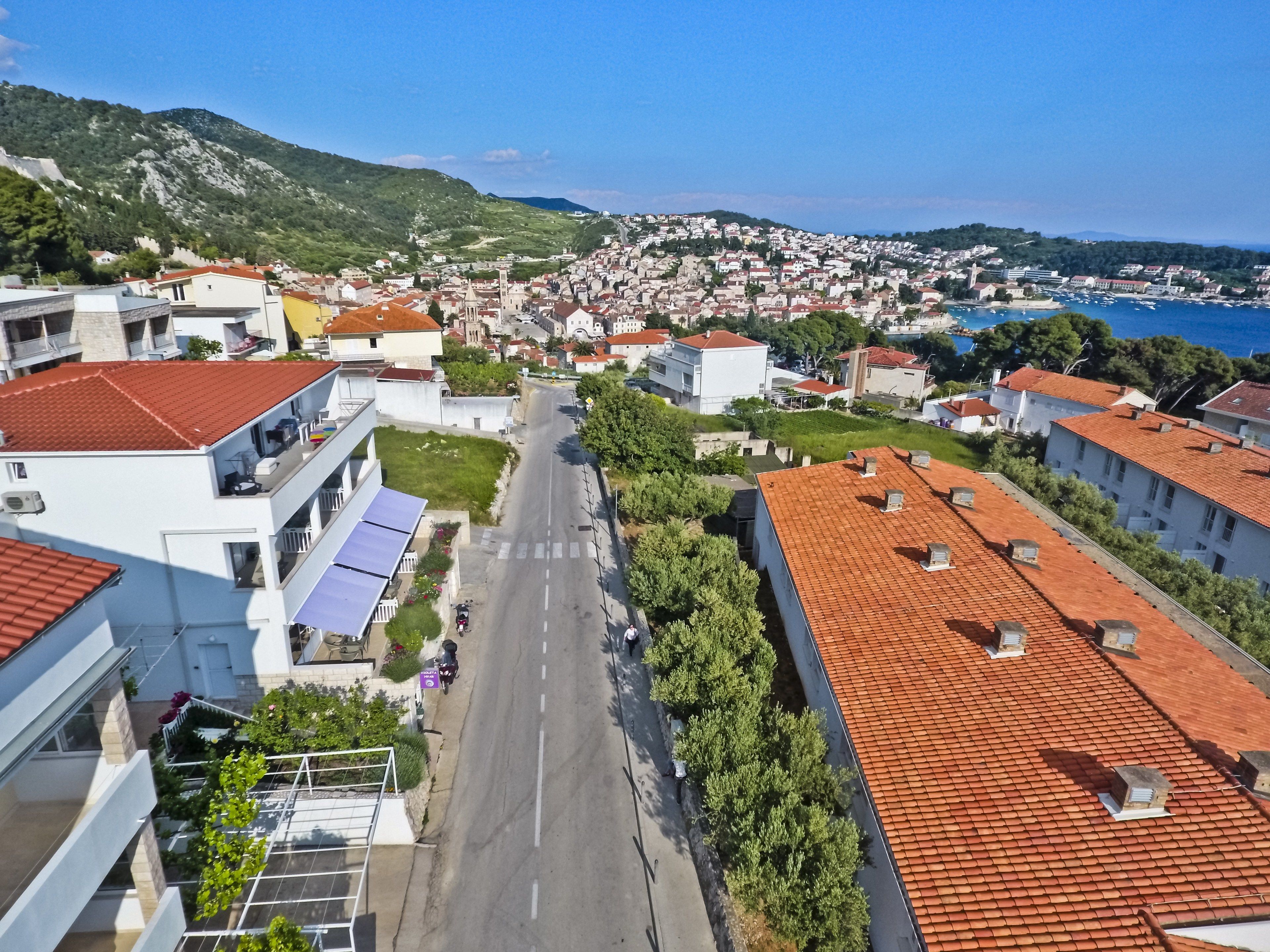 Un quartiere residenziale con una strada fiancheggiata da case, alcune con tetti in tegole rosse, con sullo sfondo delle colline e un corpo d'acqua.