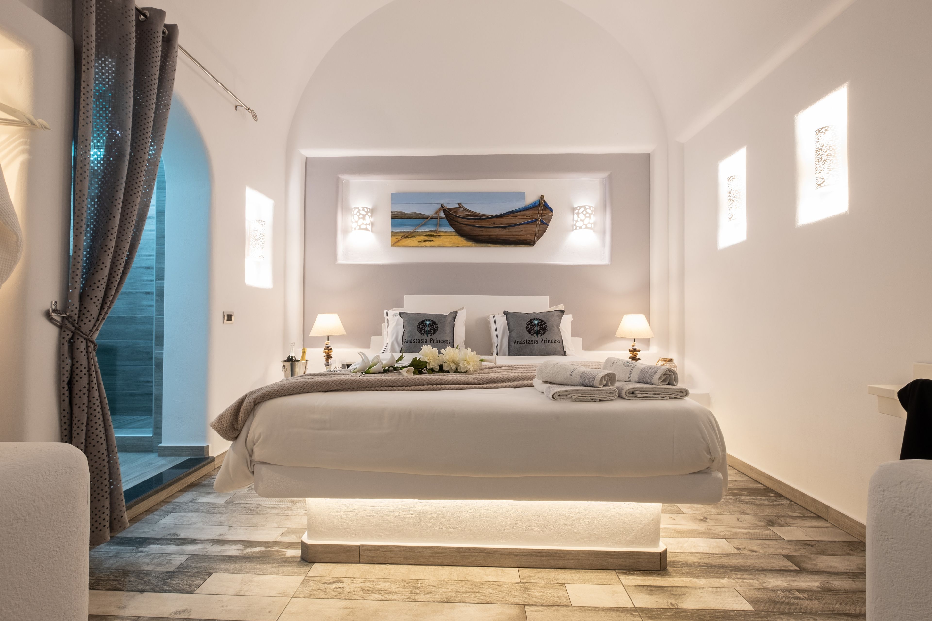 A modern bedroom with a large bed, two bedside tables with lamps, a painting of a boat on the wall, and a view of the sea through a glass door.