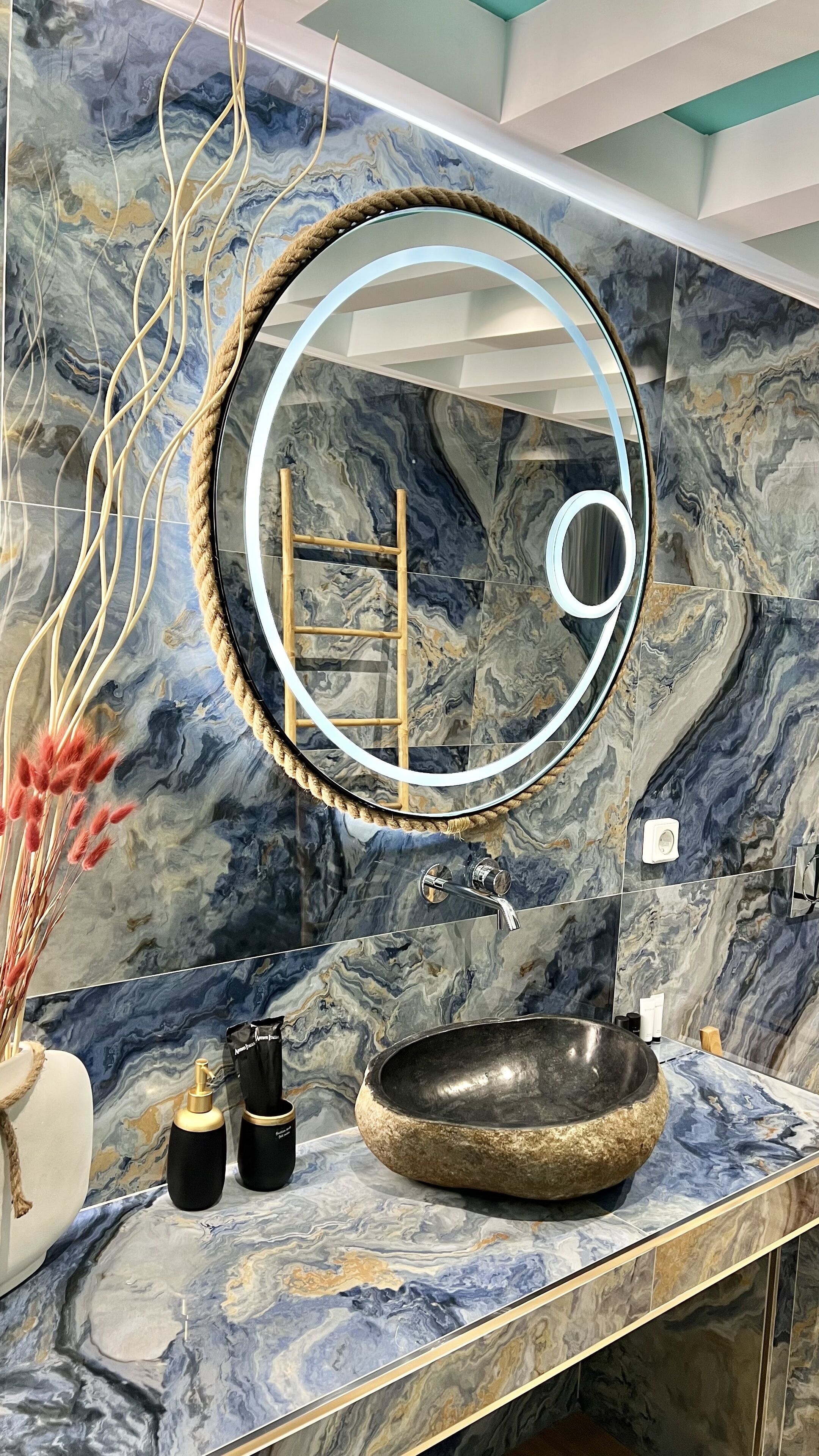 Een badkamer met een marmeren spatwand, een ronde spiegel met een touwlijst, een stenen wastafel en een klein houten krukje.