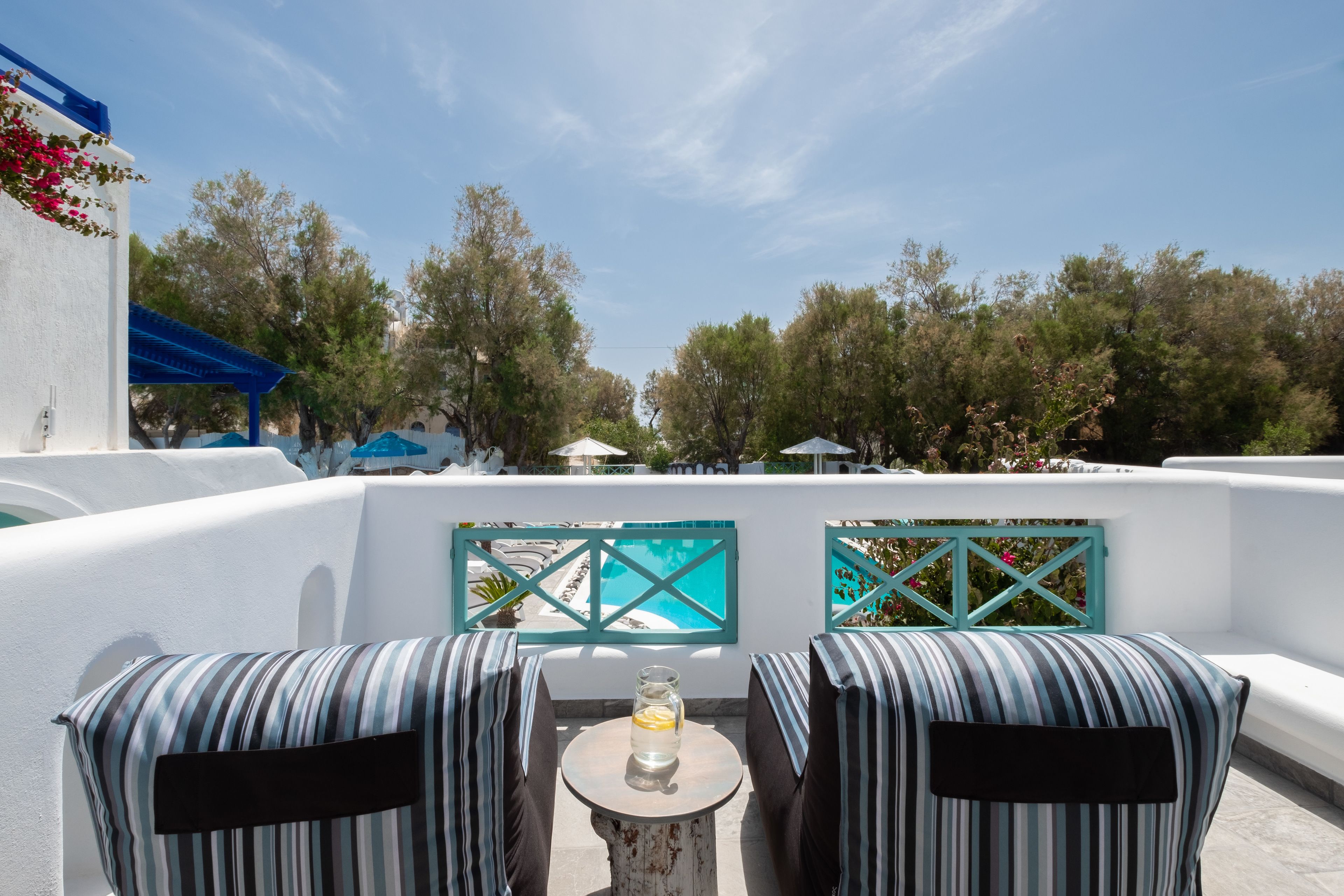 A terrace with two striped chairs, a small table with a glass of water, and a view of a pool and trees.