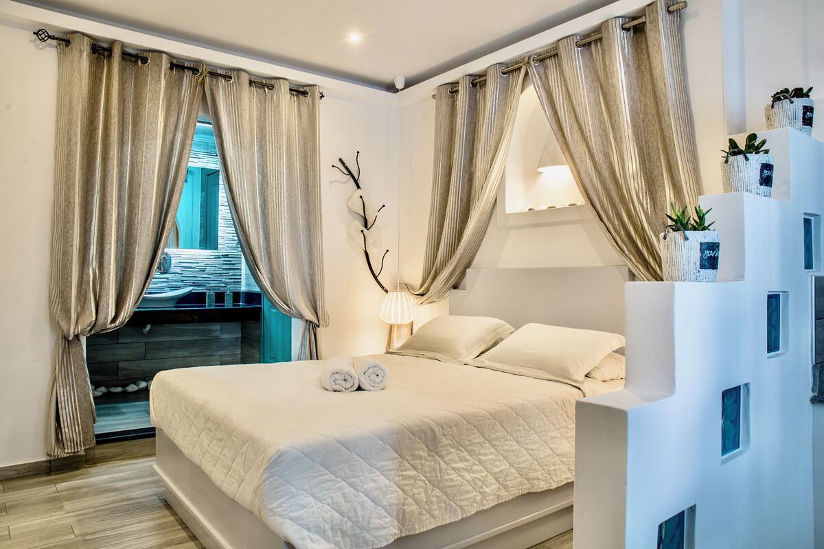 Een moderne slaapkamer met een groot bed, beige gordijnen, een bedlampje en uitzicht op een zwembad.